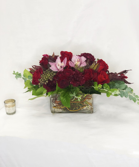 Cymbidium orchids, roses and more in shades of deep red and pink presented in a charming container.